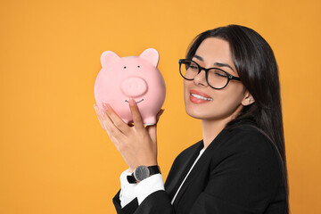 Young woman in eyeglasses with piggy bank on orange background