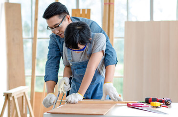 Asian professional male carpenter woodworker engineer dad in jeans outfit with safety gloves goggles helping teaching young boy son using handsaw cutting wooden stick in housing construction site