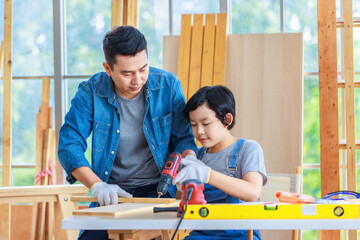 Asian cheerful male dad and son carpenter woodworker colleague in jeans outfit smiling helping using drill machine digging hole in wood stick on working workshop table in housing construction site