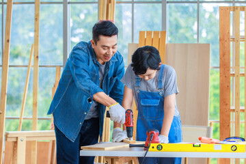 Asian cheerful male dad and son carpenter woodworker colleague in jeans outfit smiling helping using drill machine digging hole in wood stick on working workshop table in housing construction site