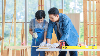 Asian happy cheerful male dad son carpenter woodworker colleague in jeans outfit with safety gloves helping using hand saw cutting wood stick on working workshop table in housing construction site