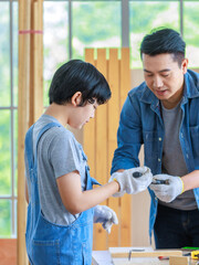 Obraz premium Asian cheerful male dad son carpenter woodworker colleague in jeans outfit with safety gloves helping using pencil drawing writing blueprint on working workshop table in housing construction site