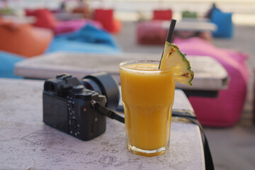 Mango juice with pineapple garnish and plastic straw in soft focus put alongside with a black mirrorless camera waiting for sunset in a beach in Bali, Indonesia.