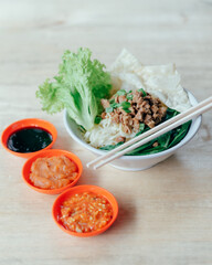 A bowl of Indonesian mie ayam or chicken noodles served with lettuce, leeks, and pangsit toppings put on bright wooden background