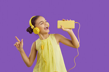 Cheerful preteen girl in headphones connected to mobile phone listens to music and sings along....