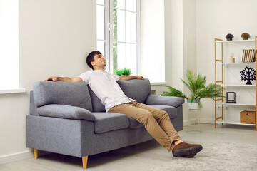 Satisfied calm young man resting after hard day's work sitting on comfortable sofa in living room at home. Smiling man enjoys lazy rest with his arms spread out on back of sofa. Rest concept.