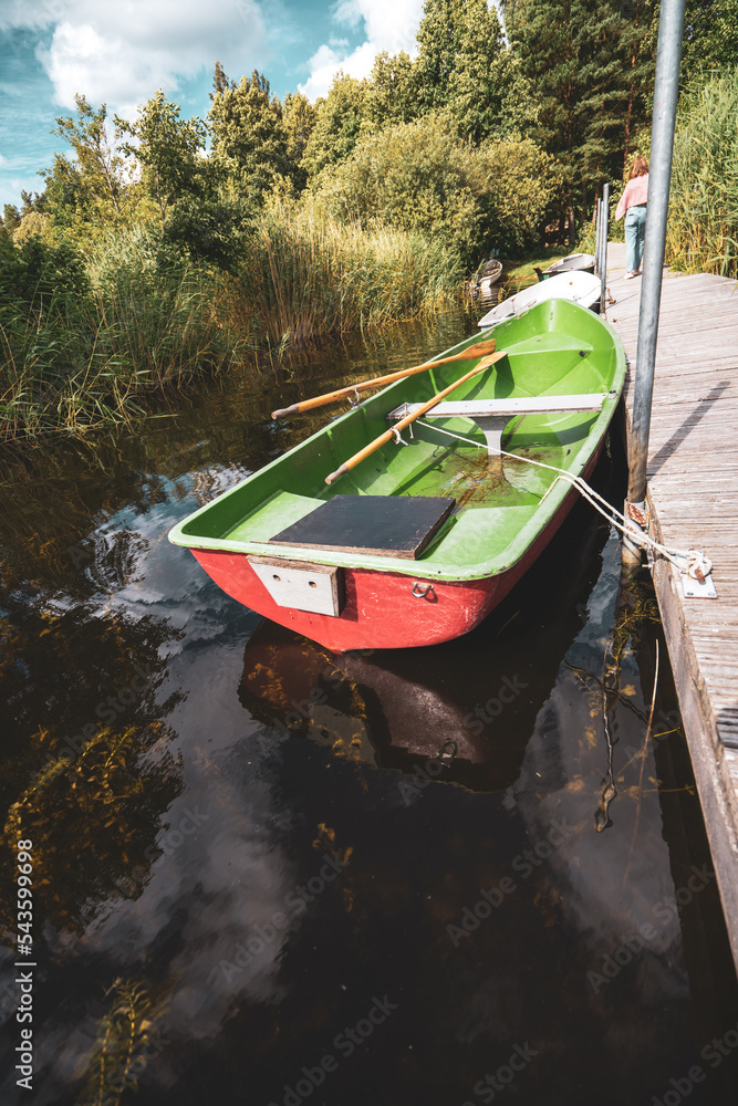 Poster a small green and red boat tied to a dock on a lake with trees in the background and a blue sky with