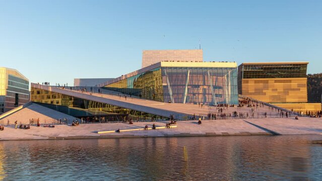 Oslo opera house hyper-lapse with people walking in the afternoon light, on a sunny day under clear blue sky. Time-lapse of the famous landmark in the capital of Norway, with water in the foreground.