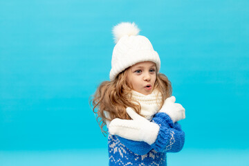 a little girl child froze on a blue background in a winter hat and sweater with mittens, space for text