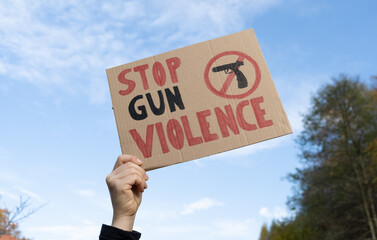 Hand holding a sign with slogan Stop Gun Violence and black pistol strikethrough. Woman with placard at protest rally demonstration strike to ban weapons and end shooting. - obrazy, fototapety, plakaty