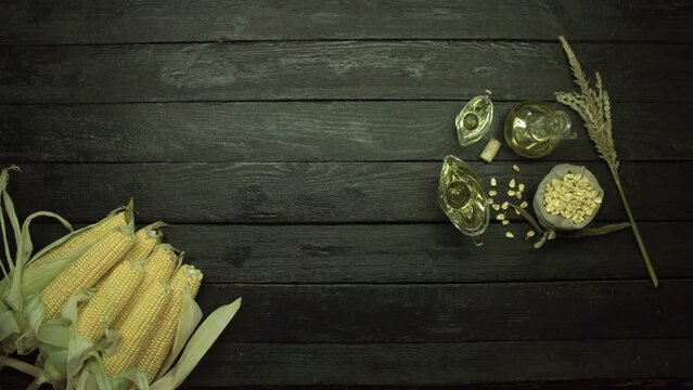 On a black wooden table is an armful of corn on the cob, as well as corn oil in various dishes, corn kernels and the top of the stem .Camera movement over the table from right to left. Flat lay.