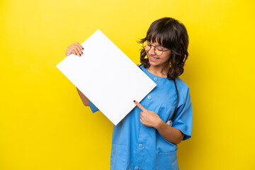 Young nurse doctor woman isolated on yellow background holding an empty placard with happy expression and pointing it
