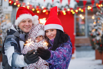 Family with sparklers in red hats and caps in Christmas decoration lightes.