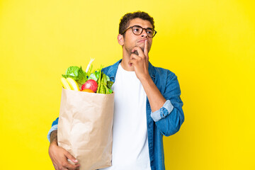 Young Brazilian man holding a grocery shopping bag isolated on yellow background having doubts...