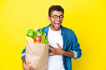 Young Brazilian man holding a grocery shopping bag isolated on yellow background smiling a lot