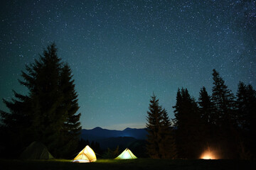 Bright illuminated tourist tents near glowing bonfire on camping site in dark mountain woods under...