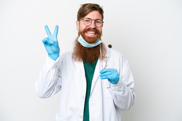 Dentist reddish man holding tools isolated on white background smiling and showing victory sign