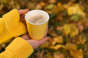 Female hands hold a yellow cup with a coffee drink, against the background of yellow leaves, autumn concept