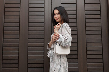 Fototapeta premium Fashionable beautiful girl in a stylish vintage summer dress with a floral print with a white trendy handbag stands near a brown wooden door