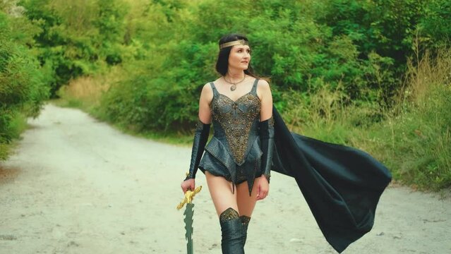 Portrait Fantasy woman viking princess warrior walks down road. girl old style sexy valkyrie holding sword in hand. Black Leather costume armor cloak flies in wind. green nature summer. art video 4k