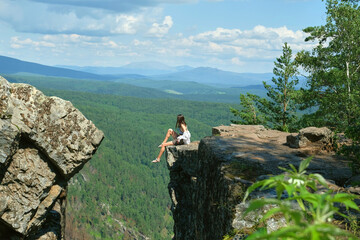A girl with long hair sits on the edge of a cliff. Landscape, valley, mountains, summer, forest, river. Panorama, height.