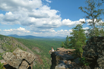 A girl with long hair sits on the edge of a cliff. Landscape, valley, mountains, summer, forest, river. Panorama, height.