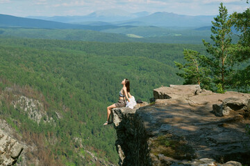 A girl with long hair sits on the edge of a cliff. Landscape, valley, mountains, summer, forest, river. Panorama, height.