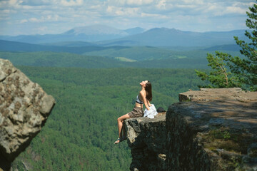 A girl with long hair sits on the edge of a cliff. Landscape, valley, mountains, summer, forest, river. Panorama, height.