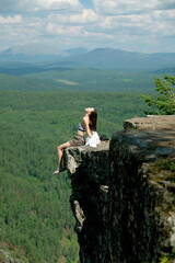 A girl with long hair sits on the edge of a cliff. Landscape, valley, mountains, summer, forest, river. Panorama, height.