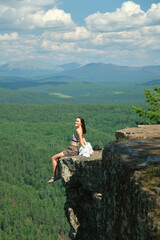 A girl with long hair sits on the edge of a cliff. Landscape, valley, mountains, summer, forest, river. Panorama, height.