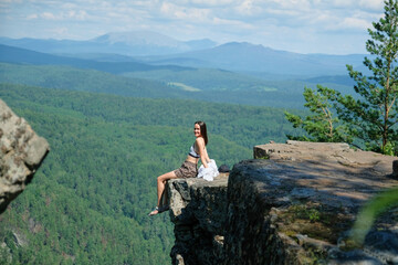 A girl with long hair sits on the edge of a cliff. Landscape, valley, mountains, summer, forest, river. Panorama, height.