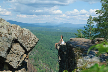 A girl with long hair sits on the edge of a cliff. Landscape, valley, mountains, summer, forest, river. Panorama, height.