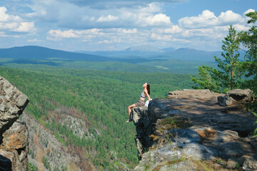A girl with long hair sits on the edge of a cliff. Landscape, valley, mountains, summer, forest, river. Panorama, height.