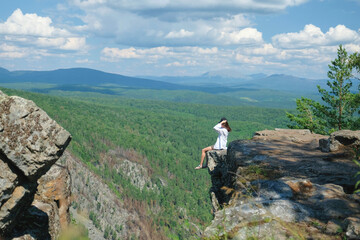 A girl with long hair sits on the edge of a cliff. Landscape, valley, mountains, summer, forest, river. Panorama, height.