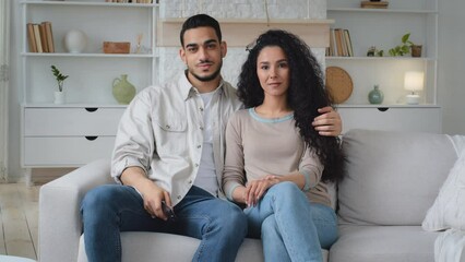 Hispanic Indian man husband loving boyfriend and Caucasian woman wife girlfriend sitting together on home couch switching TV channels with remote control watching television choosing TV program movie