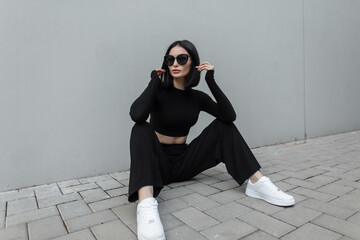 Fashionable young urban woman with black trendy sunglasses in fashion black stylish sports outfit with white sneakers sits on the street near a modern gray building - obrazy, fototapety, plakaty