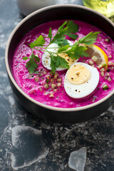 Close-up of cold beetroot soup topped with boiled egg and fresh greens, vertical shot on a dark-brown granite background