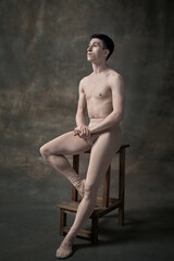Portrait of young handsome man, ballet dancer in pastel, nude color clothes posing on chair over...