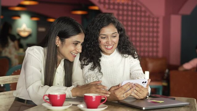  Two Indian business girl holding smart phone watching funny videos ,movie or social media content while siting on chair at the cafe.Pretty Asian woman looking at mobile screen and drinking coffee.  