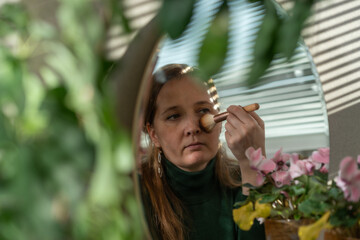 Caucasian middle aged woman uses make up brush in front of mirror. Candid lifestyle