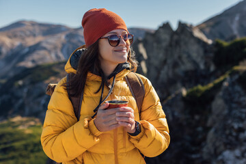 Portrait of a happy woman hiker in yellow down jacket standing with cup f tea on the slope of...