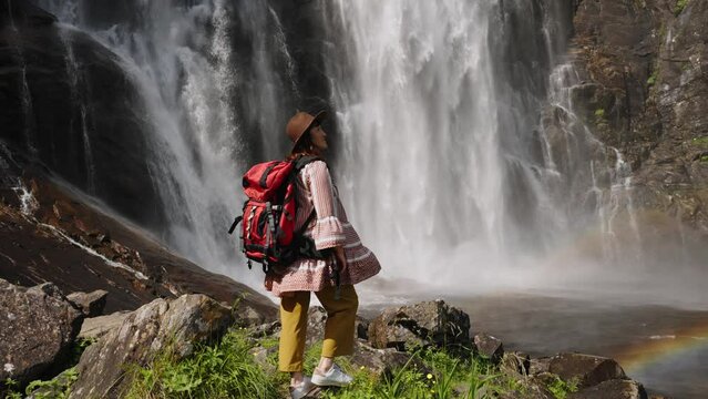 young girl standing with backpack observing a forest landscape with waterfall. Active turism norway