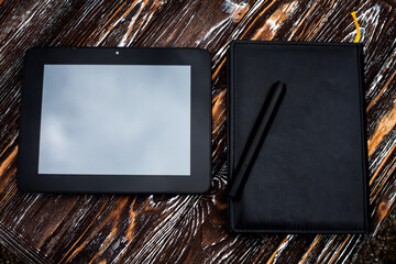A black tablet and notebook, with a leather cover, and a pen, lie on a dark wooden textured surface.