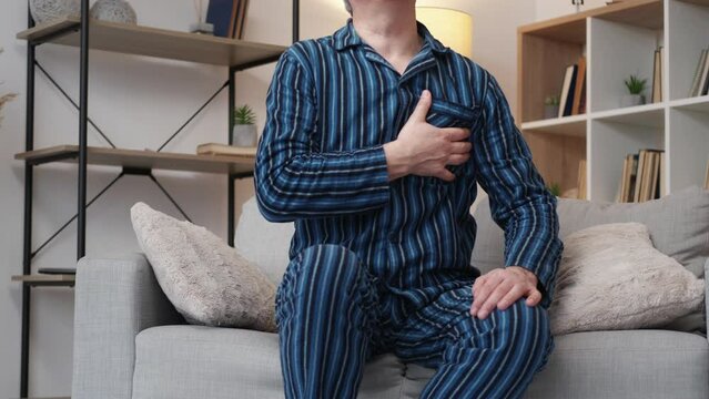 Heart problem. Aged man. Stressed disease. Unrecognizable elder male in pajamas suffering from pain in chest sitting sofa in light room interior.