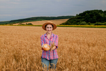 A female farmer in a straw hat and checkered shirt holds fragrant bread and ears of in her hands on a ripe wheat field. The smell of freshly baked bread