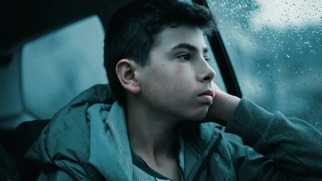 Depressed teen boy seated in car backseat looking out window during rainy day. Sad kid feeling boredom on road