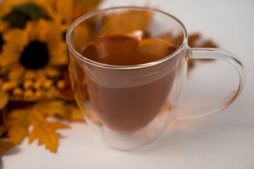 Herzförmige Tasse aus Glas mit Tee gefüllt auf weißem Hintergrund, Blick von schräg oben,  Laub und kleine Sonnenblumen als Dekoration in der linken oberen Ecke