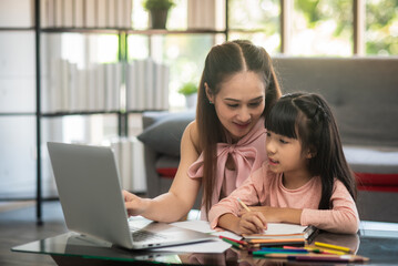 Obraz premium Asian beautiful mother pleased to see elementary school level adorable girl sit online classes In living room and take notes of the teacher's lectures via video call with a laptop. online home school