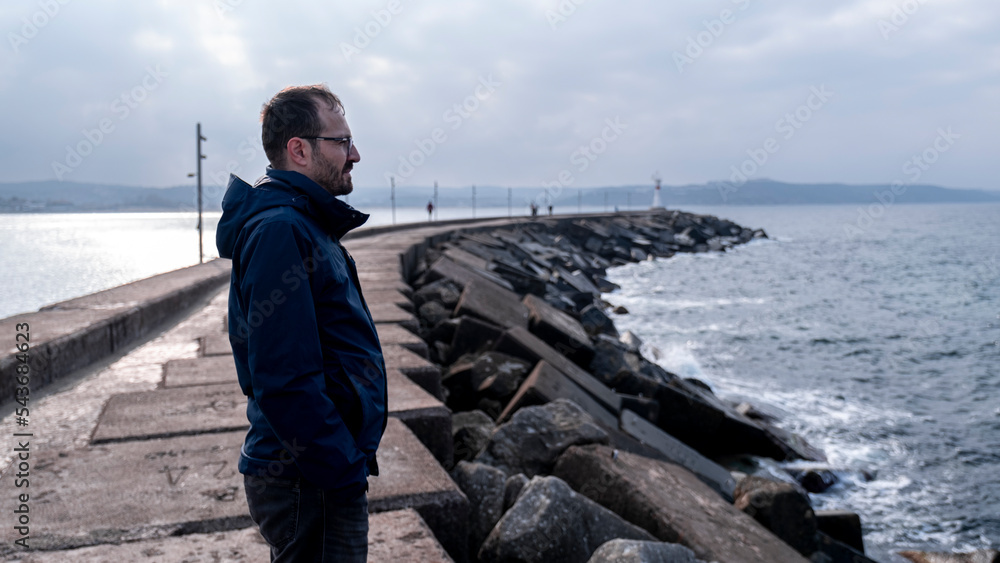 Wall mural lonely man watching open sea in harbor, thoughtful gaze, dark clouds and sea, winter weather, image 