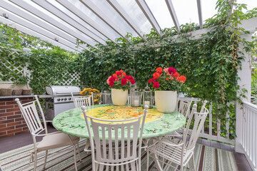 A beautiful terrace with a gazebo covered with ivy. White garden furniture and red flowers on the...